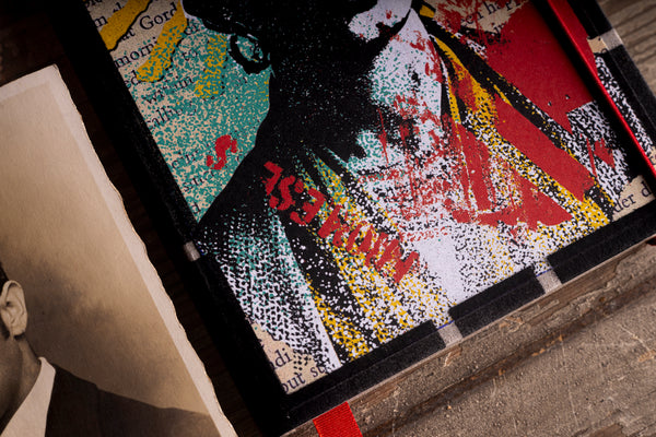 Detail of the lower part of the "Frida – the one and only" notebook cover, with layered red, yellow and black screen-print texture, a red stamped-style word, the black checkered border and a red ribbon bookmark, on a wooden surface beside a sepia photograph.