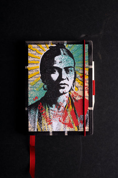 Handmade notebook "Frida – the one and only" with a screen-printed pop-art portrait cover in black, teal, yellow and red, set over torn book-page text. It has a black textured border, a red elastic closure, a red ribbon bookmark and a pen loop, shown on a black background.