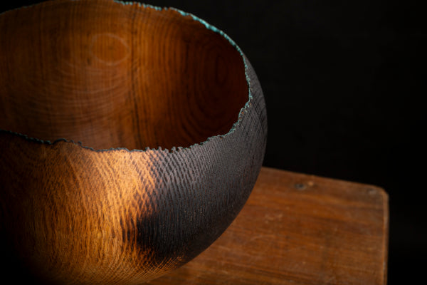 A close-up of a handcrafted wooden bowl with a rough, raw-edge rim, resting on a wooden surface against a dark background. Created by maalstrom.studio
