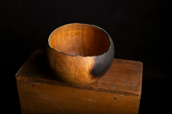 A rustic handcrafted wooden bowl with a dark charred edge, displayed on a wooden crate against a solid black background.