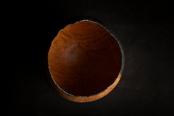 A top-down view of a handcrafted wooden bowl with a raw, irregularly edged surface, showcasing a rich warm brown wood grain against a solid black background.
