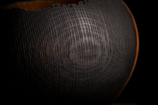 Close-up of a dark textured wooden bowl with concentric circular patterns and a warm orange-brown rim, set against a solid black background. Made from red oak.