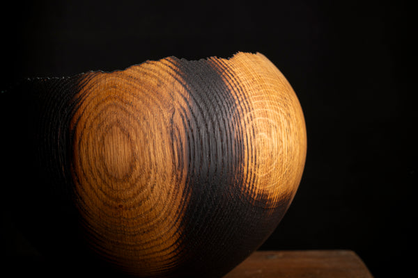A close-up of a sculptural wooden bowl with a textured, burnt-black finish on one side and warm golden-brown wood grain on the other, set against a solid black background.