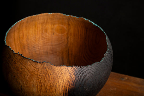 A close-up of a handcrafted wooden bowl with a rustic, unevenly broken rim, showcasing rich warm brown wood grain and a dark charred edge, set against a dark background.
