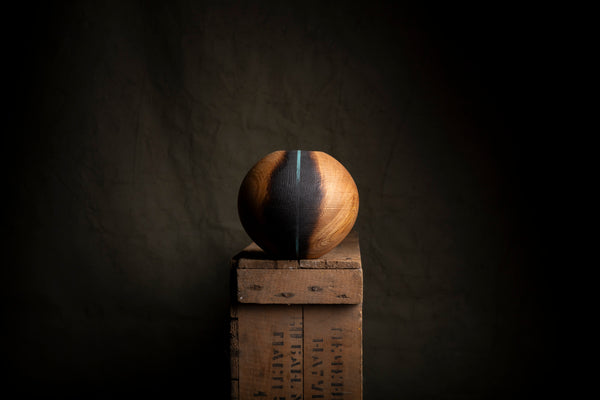 A spherical wooden hollow form with a vertical black-burned fissure, displayed on top of rustic wooden crates against a dark textured background.