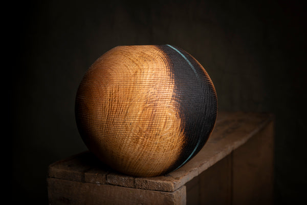 A spherical wooden hollow form with a textured, flame-burned finish in shades of brown and black, displayed on a rustic wooden block against a dark background.