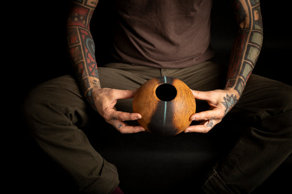 A person with tattooed arms holds a carved wooden vessel made from red oak with a circular hole in the center, set against a solid black background.