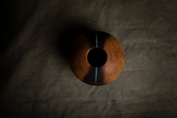 A handcrafted wooden hollow form with a dark center opening, featuring a warm brown wood grain exterior accented by two vertical light blue stripes, set against a dark textured fabric background.