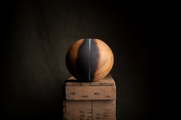 A spherical wooden object from red oak with a dark center and warm brown wood grain edges, displayed on top of stacked rustic wooden crates against a dark studio background.