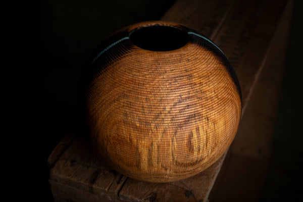 A round wooden hollow form with a textured, carved surface and a dark opening, resting on a rustic wooden block against a dark background. This object is handturned from red oak.