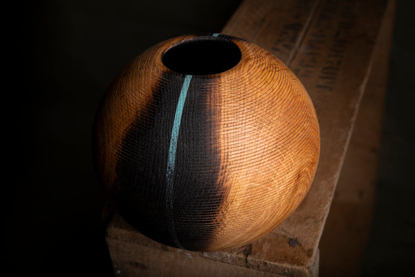 A spherical wooden hollow form with a textured grain finish, featuring a vertical black charred stripe down the center and a small turquoise accent line near the top, resting on a rustic wooden crate against a dark background.