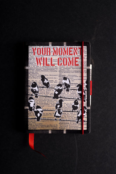 Handmade notebook "Birds on a Wire" with a screen-printed cover of black and white birds perched on wires over a German dictionary page, under the red stencil words "YOUR MOMENT WILL COME". It has a black textured border, a red elastic closure, a red ribbon bookmark and a pen loop, on a black background.
