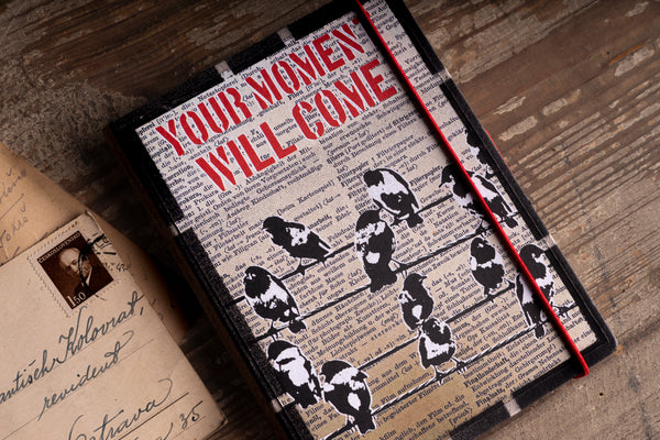 Angled close-up of the "Birds on a Wire" handmade notebook cover, showing black and white screen-printed birds perched on wires over German dictionary text, with the red stencil words "YOUR MOMENT WILL COME" and a red elastic band, beside an old postcard with a vintage stamp on weathered wood.