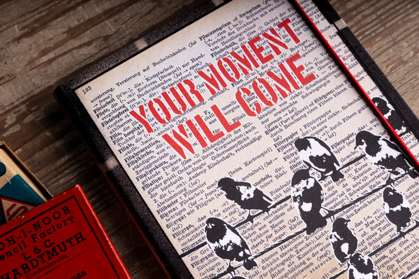 A notebook featuring the phrase 'YOUR MOMENT WILL COME' in bold red text over a vintage book page background with black bird silhouettes. The frame is shown at an angle on a wooden surface next to another red box.