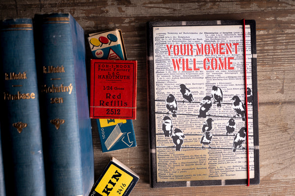 A hardcover journal with a vintage dictionary cover and the words 'YOUR MOMENT WILL COME' in red, styled on a wooden table next to old blue books and matches.