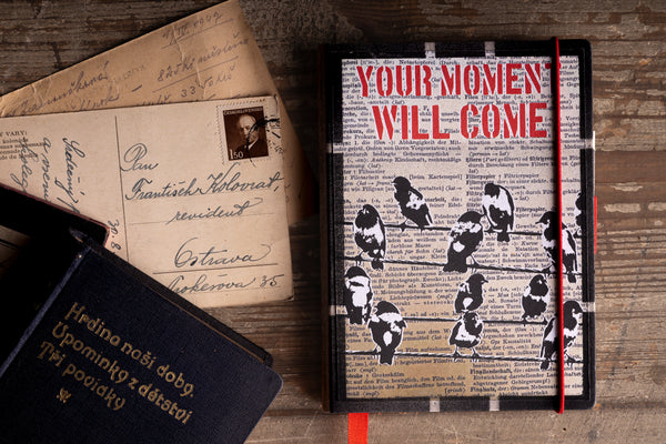 Handmade notebook "Birds on a Wire" with a screen-printed cover of black and white birds on wires and the red stencil text "YOUR MOMENT WILL COME", beside vintage handwritten postcards and an old dark blue book with gold lettering on a rustic wooden table.