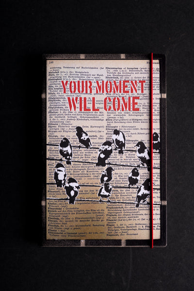 A hardcover journal featuring a vintage dictionary page background with black birds perched on wires and the bold red text 'YOUR MOMENT WILL COME'. It has a black cover, a red elastic closure, and is photographed flat on a dark background.