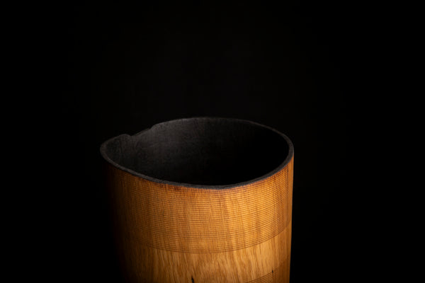 A close-up of a cylindrical wooden container or vessel with a warm honey-brown wood grain exterior and a dark interior, set against a solid black background.