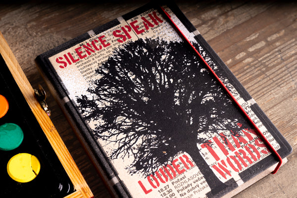 A hardcover diary with a black tree silhouette on a textured beige cover, featuring a red elastic closure, placed on a wooden surface next to an artist's paint palette.