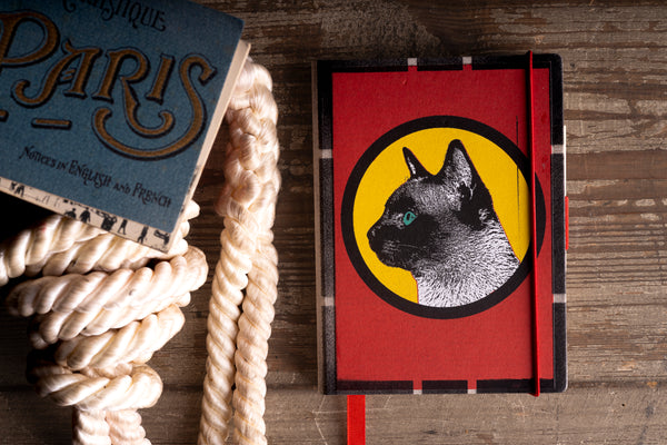A red hardcover notebook with a black and white cat illustration inside a yellow circle on the cover, styled on a rustic wooden table next to a vintage Paris book and a coiled rope.