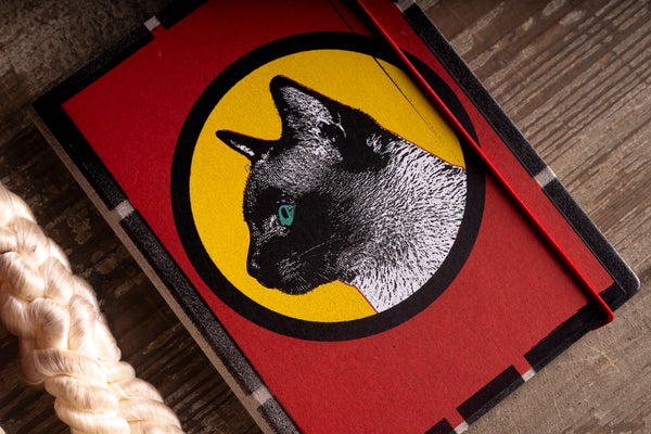 A red notebook or journal with a black and white cat illustration inside a yellow circle on the cover, photographed on a wooden surface with a braided rope prop at the lower left.