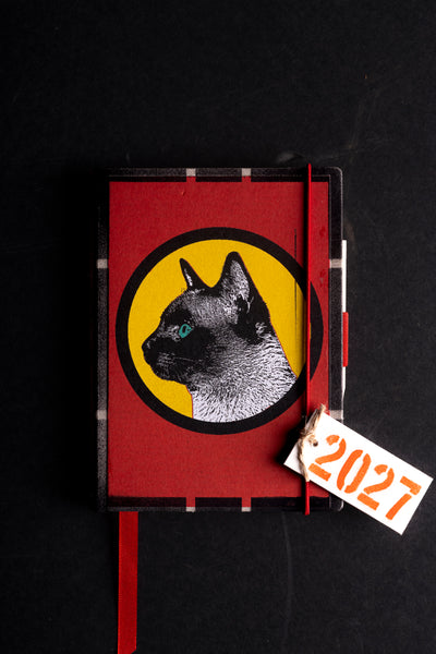 A red hardcover journal featuring a graphic of a black cat inside a yellow circle on the cover, with a red ribbon bookmark and a white paper tag reading '2027' on a black background.