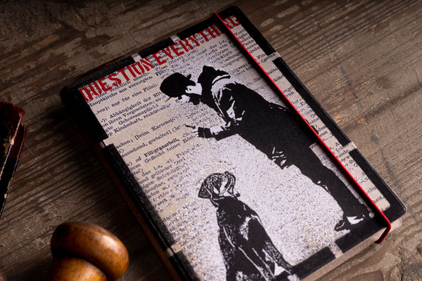 A vintage-style hardcover notebook with a black and white graphic print of a man and dog, featuring red text and a red elastic closure, resting on a rustic wooden table.