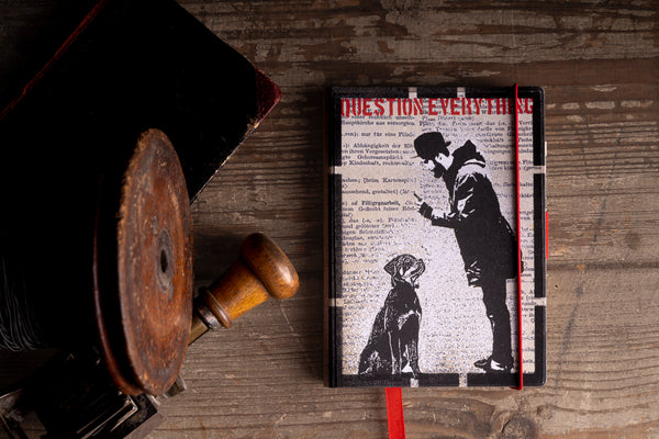 A hardcover notebook with a black and white graphic cover featuring a silhouette of a man and a dog, styled on a rustic wooden table.