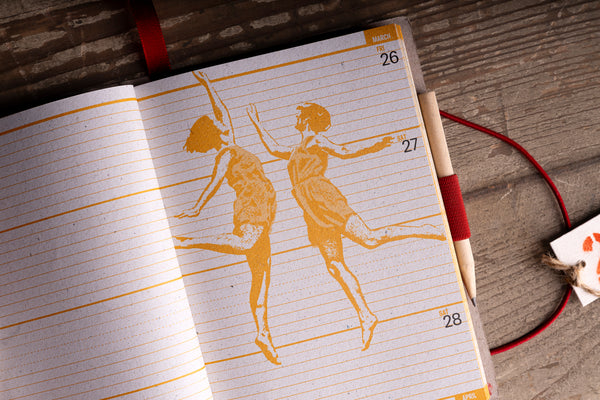 An open lined notebook or planner on a rustic wooden surface, showing two orange dancer silhouettes printed across the pages and calendar dates 26, 27, and 28. A red ribbon bookmark and a pencil are visible.