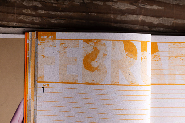 An open notebook or planner showing a February weekly layout with orange printed text and lined pages, photographed against a rustic wooden background.