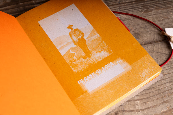 An open vintage-style book or journal with a bright orange cover and a printed sepia-toned illustration on the inner page, placed on a rustic wooden surface with a red cord visible.
