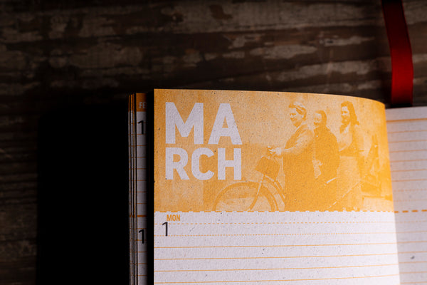 A close-up of an open planner or notebook showing a yellow-orange cover page with the word "MARCH" and a faded photo of people with a bicycle, set against a dark rustic wooden background.