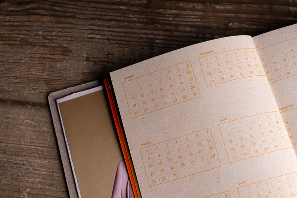 An open kraft-paper planner or calendar showing printed monthly calendar grids in orange, resting on a rustic dark wood surface next to a closed matching planner.