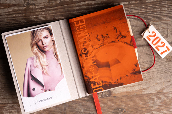 An open 2027 photo journal lies on a rustic wooden table. The left page features a printed photograph of a woman in a pink coat, while the right page is a warm orange-brown with the word 'DEADLINE' and a large circular graphic.