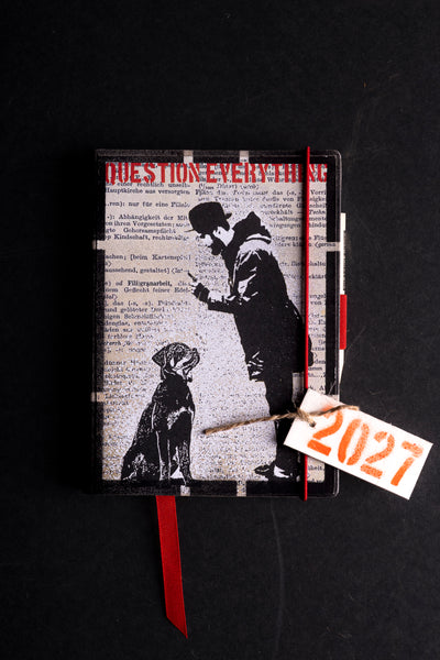 A black hardcover journal with a printed cover featuring a black-and-white illustration of a man and dog over a dictionary-style background, with red text and ribbon bookmarks, photographed on a dark surface.
