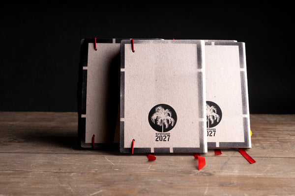 Three square paper-bound books or journals with textured off-white covers, standing upright on a wooden surface against a dark background. Each cover has a black circular palm tree logo and the year 2027 printed near the bottom, with red ribbon bookmarks visible.