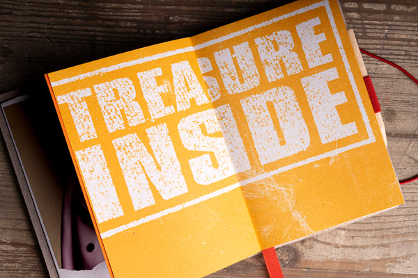 A yellow-orange notebook or journal lying open on a rustic wooden surface, with large distressed white text reading 'TREASURE INSIDE' across the cover. A red bookmark ribbon is visible, along with part of a box and a pen in the background.