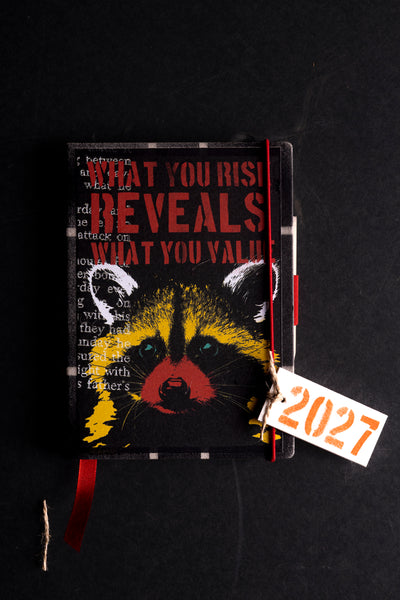 A hardcover diary with a black cover featuring a stylized raccoon face in yellow, red, and blue, with the text 'WHAT YOU RISK REVEALS WHAT YOU VALUE'. A red ribbon bookmark and a white paper tag with '2027' in orange are attached.