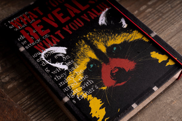 A close-up of a hardcover diary lying on a rustic wooden surface. The cover is black with a large stylized raccoon face in yellow, red, and black, overlaid with white distressed text.