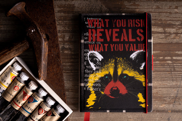 A hardcover diary with a black cover featuring a stylized raccoon face in yellow, red, and white, with the text 'WHAT YOU RISK REVEALS WHAT YOU VALUE' in red. The diary is styled on a rustic wooden table next to a paint palette and a wooden mallet.