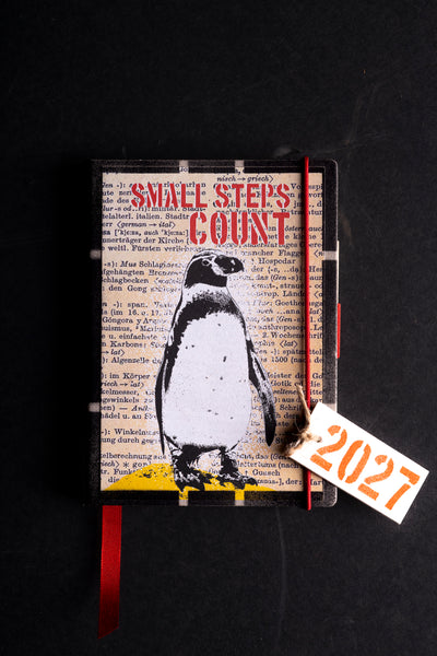 A hardcover diaryl with a penguin illustration and the words 'SMALL STEPS COUNT' on the cover, photographed on a dark background. A red elastic closure, red ribbon bookmark, and a small paper tag reading '2027' are attached.