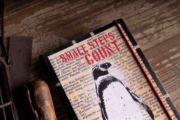 A vintage-style hardcover diary titled 'SMALL STEPS COUNT' with a black-and-white penguin illustration on the cover, placed on a rustic wooden surface with a red pencil resting along the right edge.