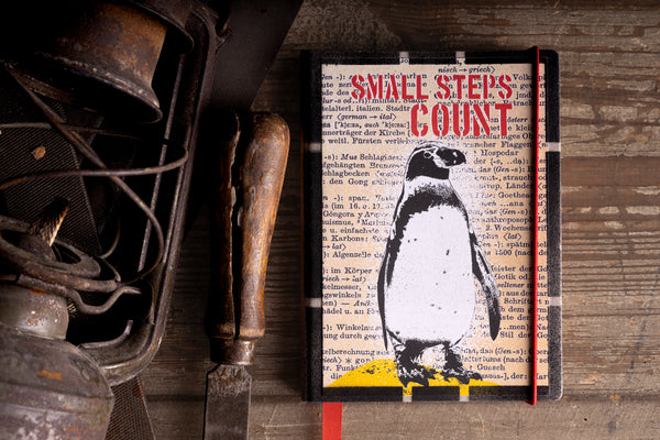 A hardcover diary featuring a black and white penguin illustration over a vintage book page background, styled on a rustic wooden surface next to an antique metal object.
