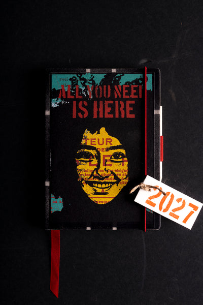 A diary with a distressed graphic design featuring a yellow face and the text 'ALL YOU NEED IS HERE' in red and yellow. A red ribbon bookmark hangs from the bottom, and a white paper tag tied with string reads '2027' in orange.