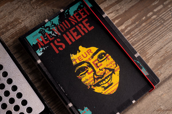A black hardcover diary with a graphic cover reading 'ALL YOU NEED IS HERE' in red and yellow over a wood-grain background.