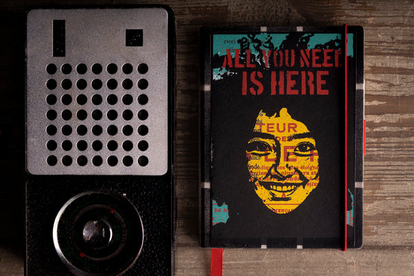 A black hardcover diary with a graphic cover featuring a yellow face and the text 'ALL YOU NEED IS HERE' in red, placed next to a vintage black radio on a rustic wooden background.