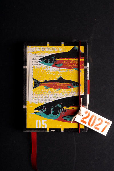A hardcover diary with a yellow book-page background and three colorful fish illustrations, photographed on a dark surface. A red elastic closure and red ribbon bookmark are visible, along with a small white tag marked 2027.
