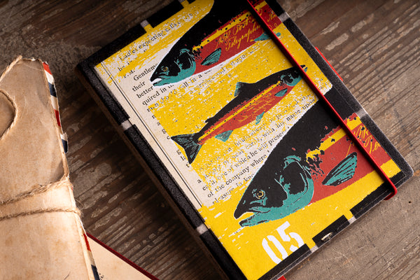 A hardcover diary with a vintage-style fish illustration on a yellow background, featuring a red elastic closure, placed on a rustic wooden surface next to a beige leather strap.