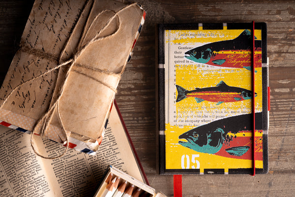 A hardcover diary with a street-style fish illustration on a yellow background, styled on a rustic wooden table next to old books and a tied leather-bound book.