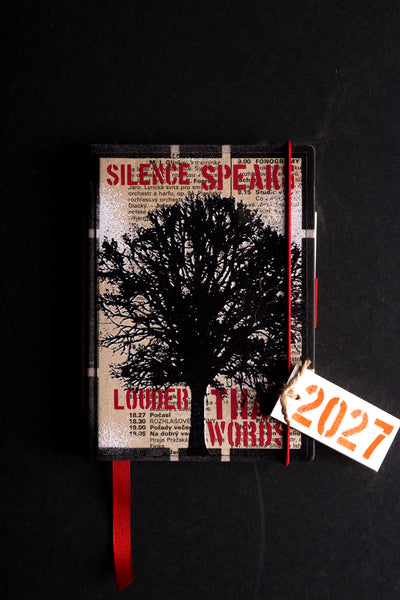 A hardcover diary featuring a black tree silhouette on a distressed beige background with red text, a red elastic closure, a red ribbon bookmark, and an attached white paper tag marked '2027'.